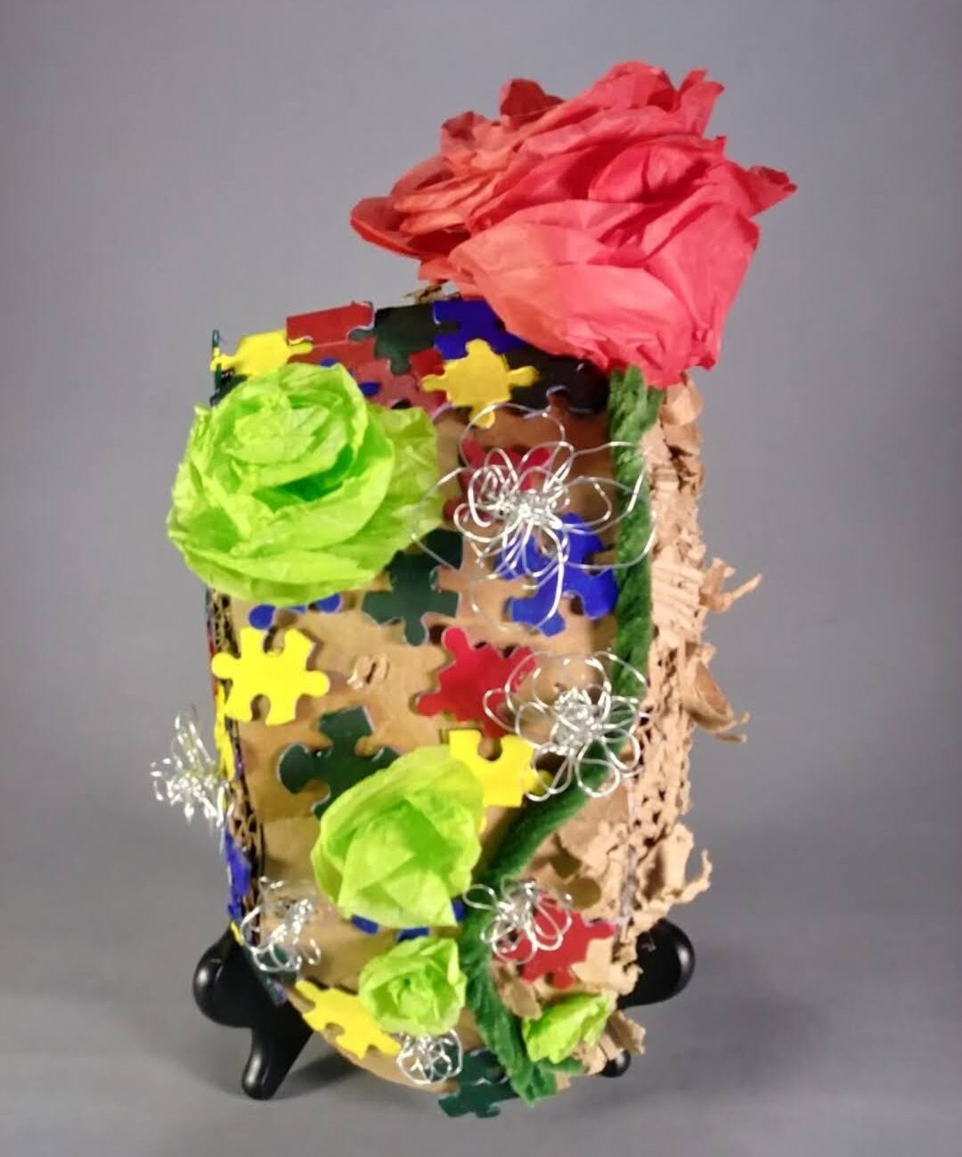 A sculpture of a mask with many various colored puzzle pieces, and a flower made of tissue paper wrapping around it.