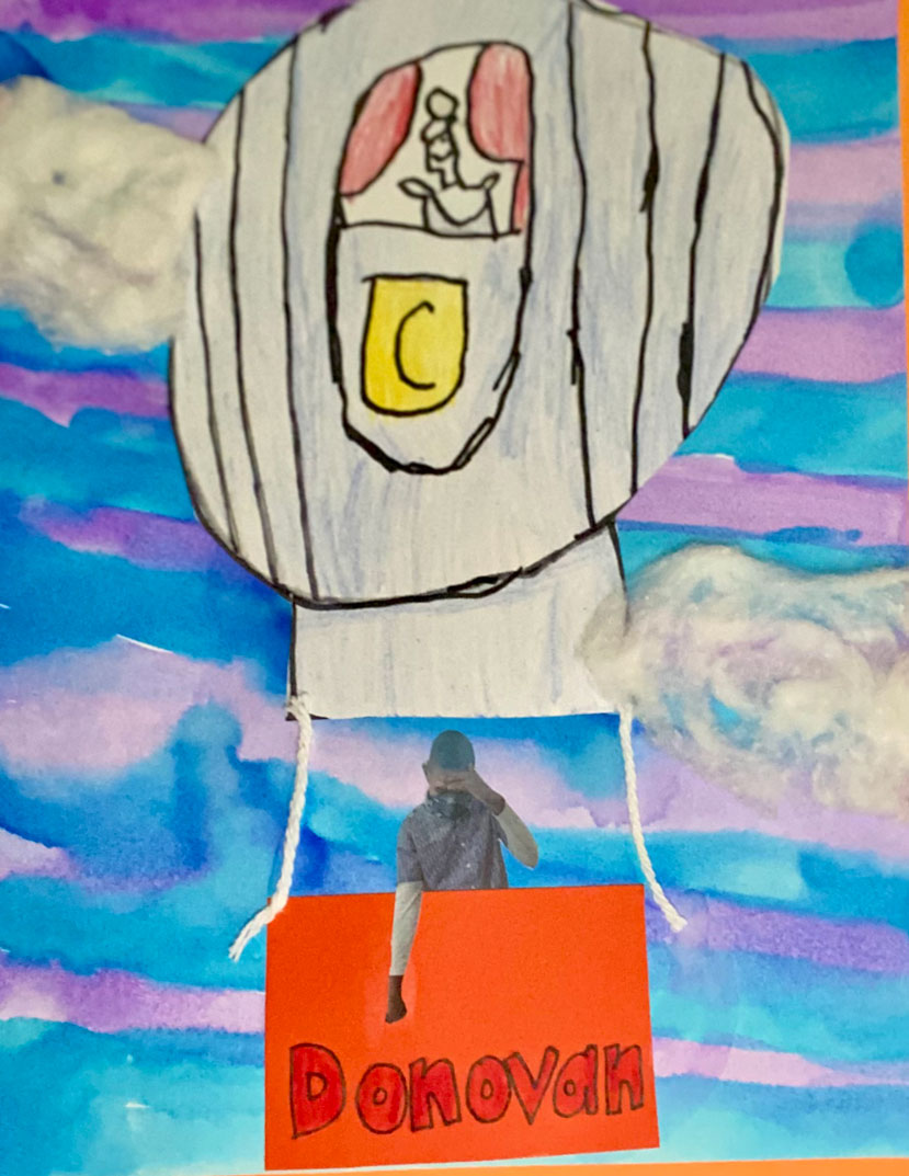 An art piece of a cut out and drawn and colored hot air balloon with a picture of a boy in the basket.