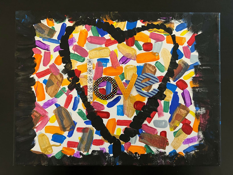 A child's art piece of a heart with the word 'love' in the middle in the foreground, and with multiple strokes of paint in many colors in the background.