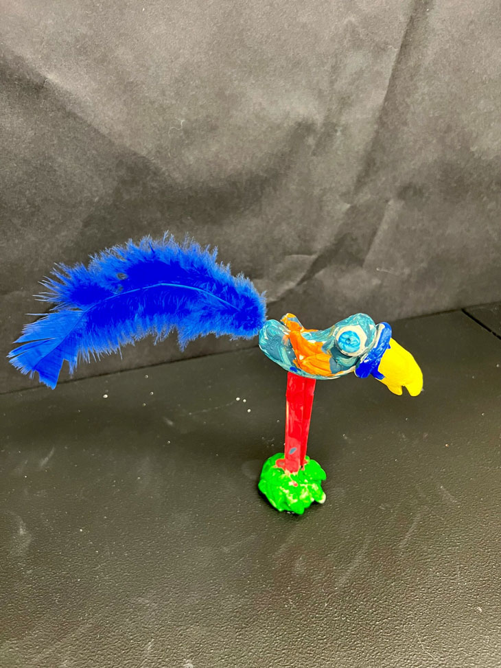 A clay sculpture of a light blue exotic bird, painted with other primary colors and has a dark blue feather.