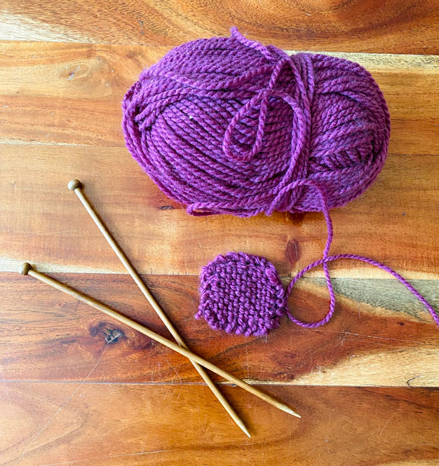 A picture of a pile of purple yarn with needles, and a small purple yarn coaster.